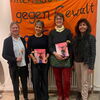 Zu sehen sind die Veranstalterinnen der literarischen Weinprobe mit einem Geschenk und Blume in der Hand vor einem orangenen Banner mit der Aufschrift "Internationaler Tag gegen Gewalt an Frauen"