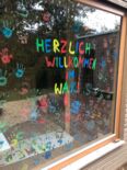 Bild des Waldkindergartens am Hoffnungshaus. Das Fenster des Kindergartens ist bunt beschriftet mit "Willkommen im WAKI" und bunte Handabdrücke der Kinder und Erzieher darunter.