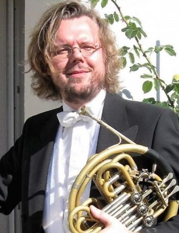Bild des Musikschul-Lehrers Martin Schilling. Er trägt eine runde Brille, einen schwarzen Anzug mit weißem Hemd und weißer Fliege. In der Hand hält er ein Horn.