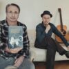 Zwei Herren in einem Wohnzimmer. Der eine hält ein Buch in der Hand, der andere sitzt auf einem Sofa, neben ihm steht eine Gitarre.